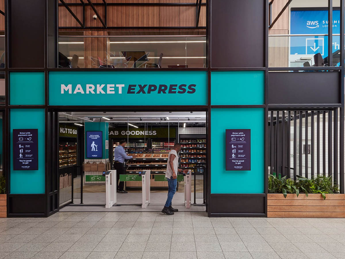 Entrance to a modern grab-and-go Market Express store, featuring automated gates and digital signage, with people entering and shelves of products visible inside.