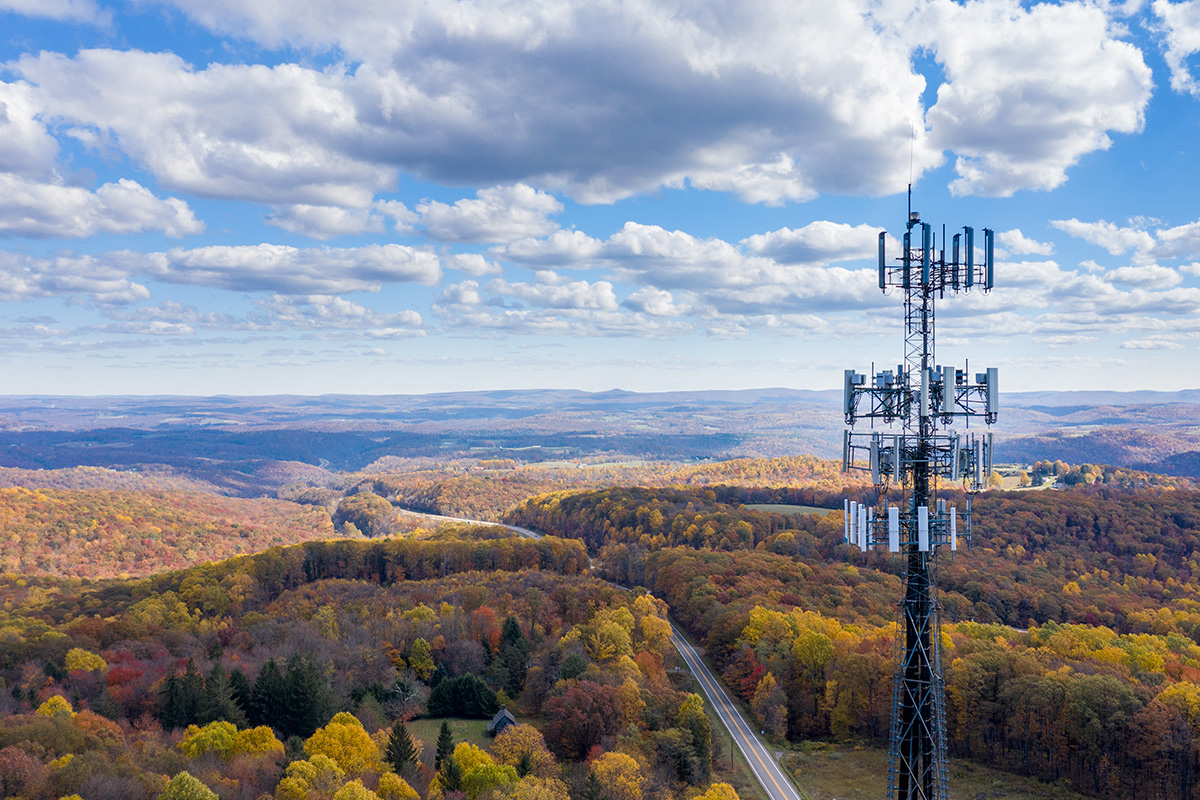 A telecommunications tower stands tall in a rural landscape with a winding road and forested hills in autumn, under a partly cloudy sky.