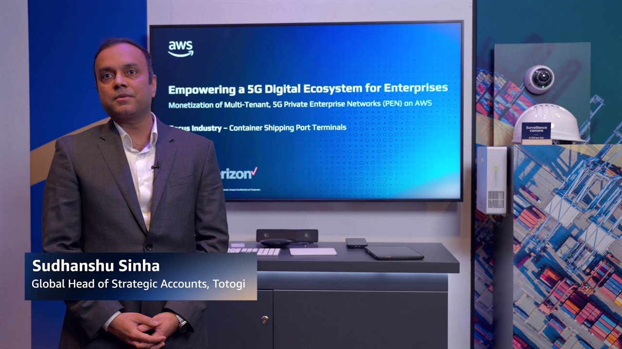 Sudhanshu Sinha, Global Head of Strategic Accounts at Totogi, stands in front of a display screen discussing monetization of multi-tenant 5G private enterprise networks on AWS for container shipping port terminals, with Verizon branding shown. The setting includes surveillance technology and digital ecosystem visuals, representing AWS 5G enterprise solutions.