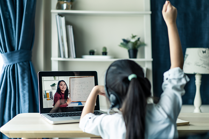 A young girl wearing headphones raises her hand while attending an online class on a laptop in a home setting.