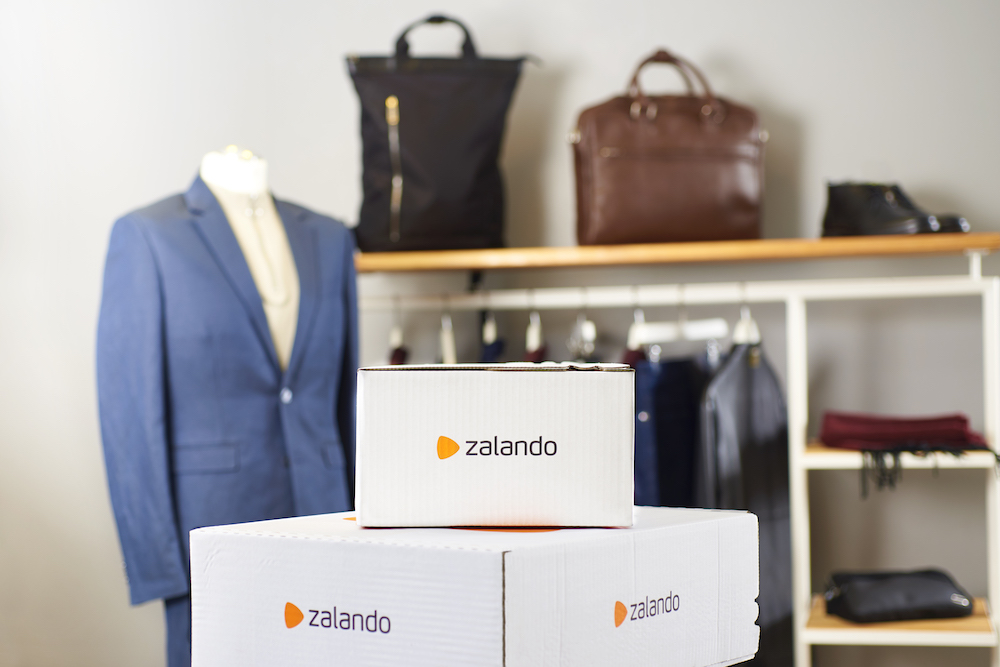 Zalando-branded boxes in a stylish showroom with a mannequin in a blue suit, bags, and clothing racks in the background.