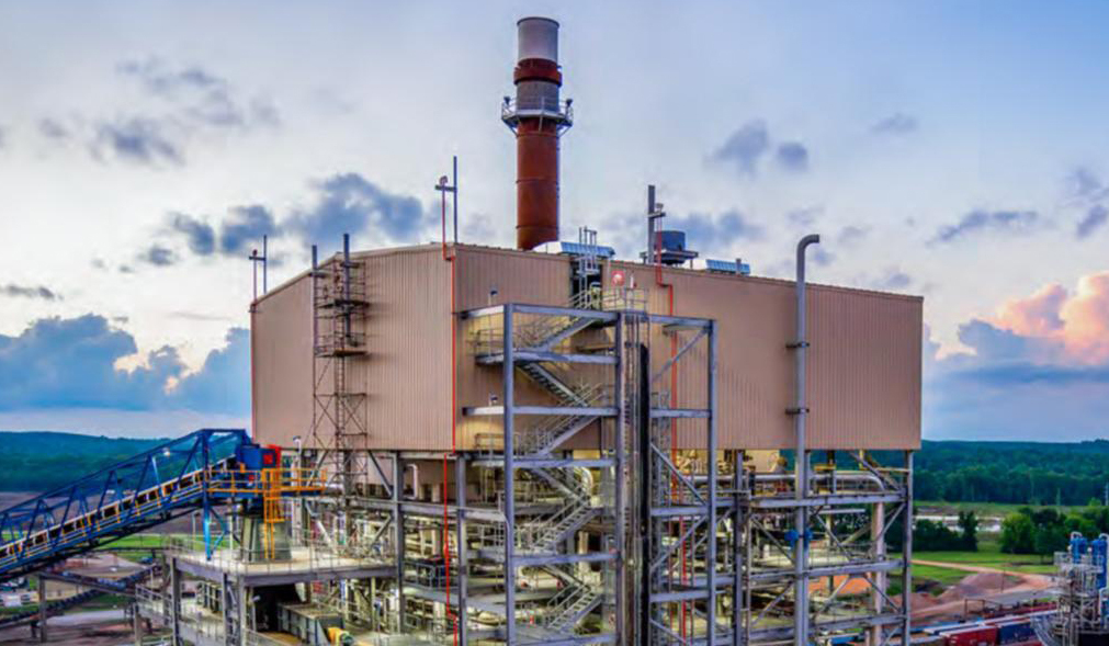 A large industrial power plant facility with metal structures, piping, and a central smokestack, situated outdoors against a scenic sky and landscape.