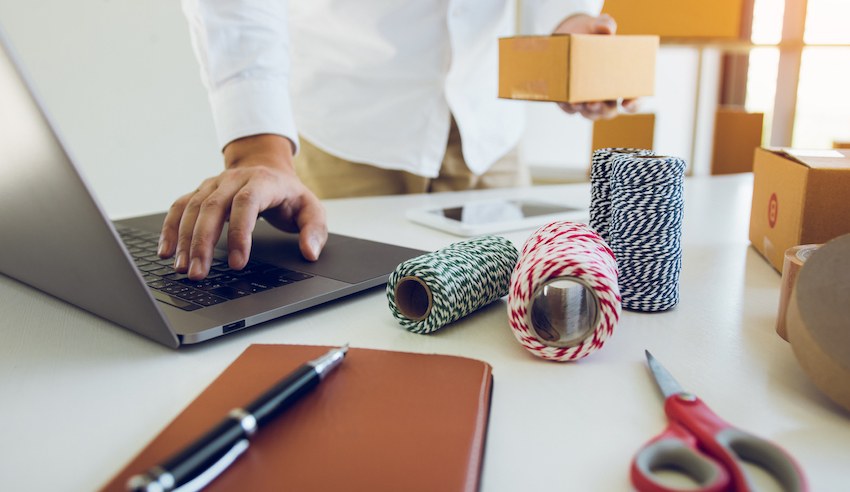 A small business owner prepares and processes online orders, using a laptop and handling packages on a desk with shipping supplies such as twine, boxes, notepad, and scissors.