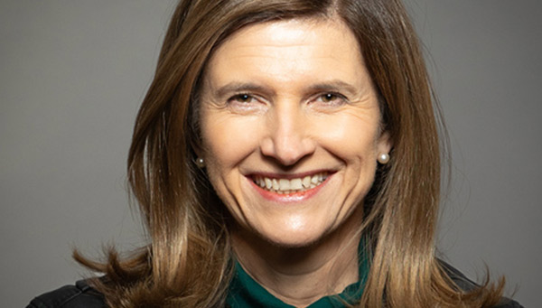 A portrait of a smiling woman with brown hair wearing a green top, set against a neutral background.