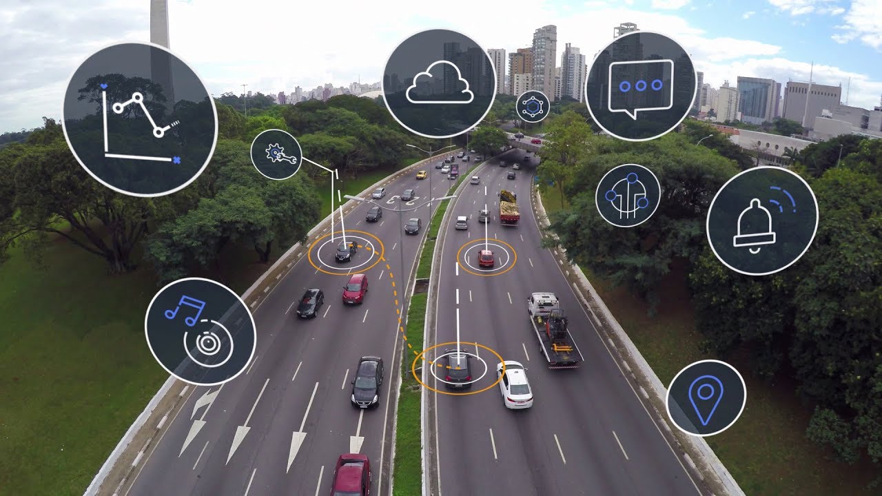 Aerial view of a multi-lane highway in an urban setting, with numerous cars and trucks driving on the road. Overlaid are icons representing cloud connectivity, data analysis, notifications, and smart technology, illustrating the concept of connected vehicles, smart cities, and intelligent transportation systems.