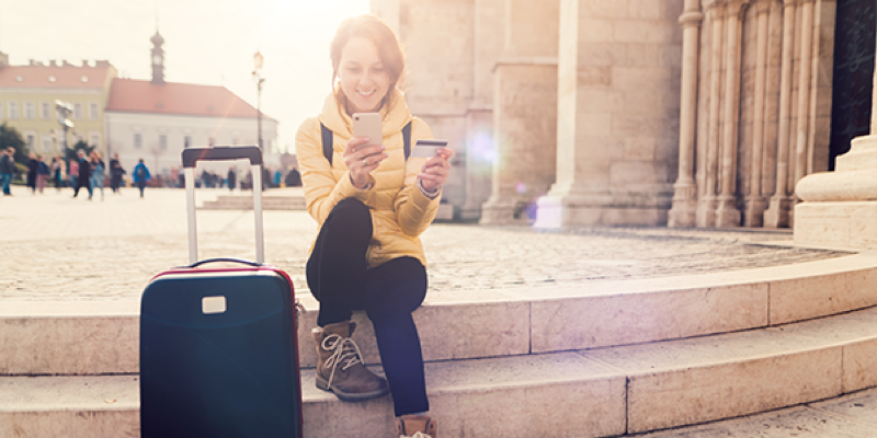 A woman traveler sits on stone steps outdoors with her luggage, smiling as she uses her phone and holds a credit card. Sunlight and historic architecture are in the background.