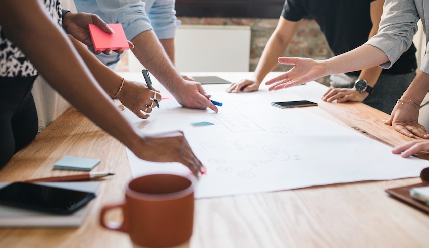 A group of people working together around a table, discussing and planning over a large sheet of paper with notes, phones, and coffee cups.