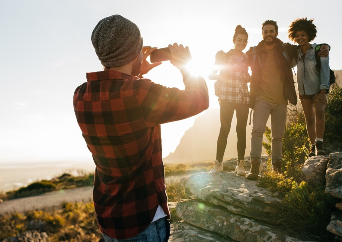 A person in a plaid shirt and beanie takes a photo of three people standing on rocks during a sunny outdoor hike.