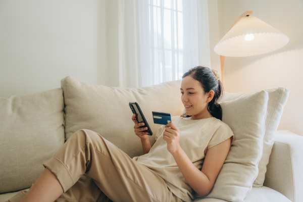 A woman relaxing on a couch using her phone and holding a credit card, representing online shopping from home.