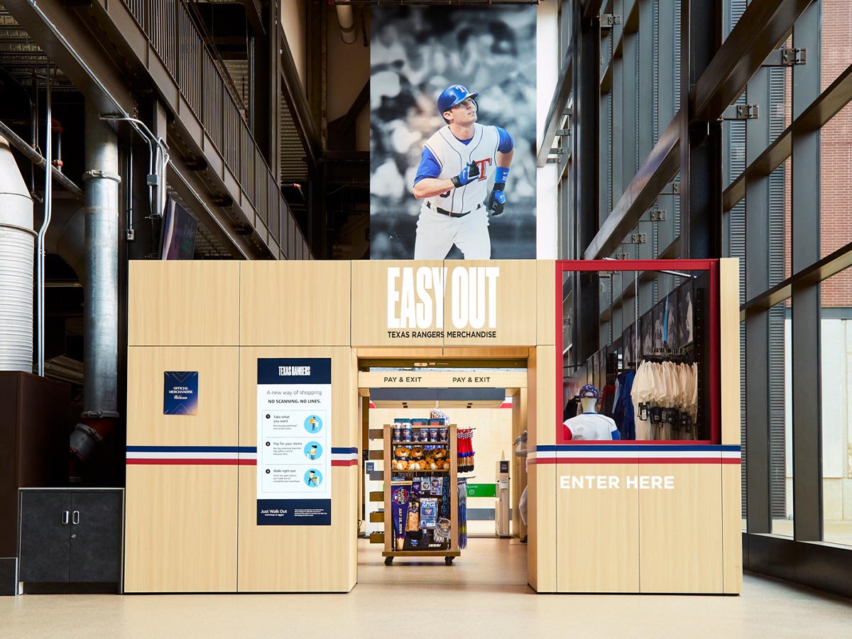 A view of the Just Walk Out Texas Rangers merchandise store located inside the Texas Rangers stadium. The store features an automated checkout experience with 'Enter Here' and 'Easy Out Texas Rangers Merchandise' signage, sports apparel, and a large image of a Texas Rangers baseball player above the entrance.