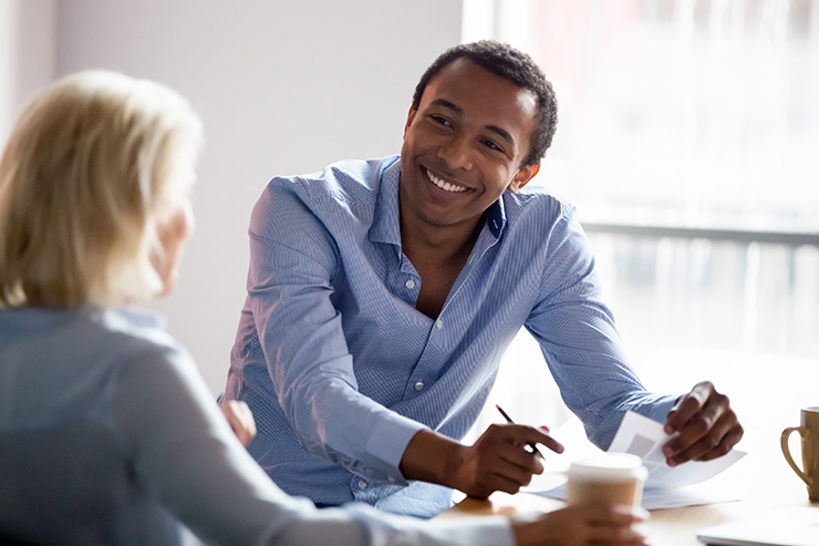 A smiling manager in a blue shirt talks with a client during a business meeting at an office table, with papers and coffee cups visible.