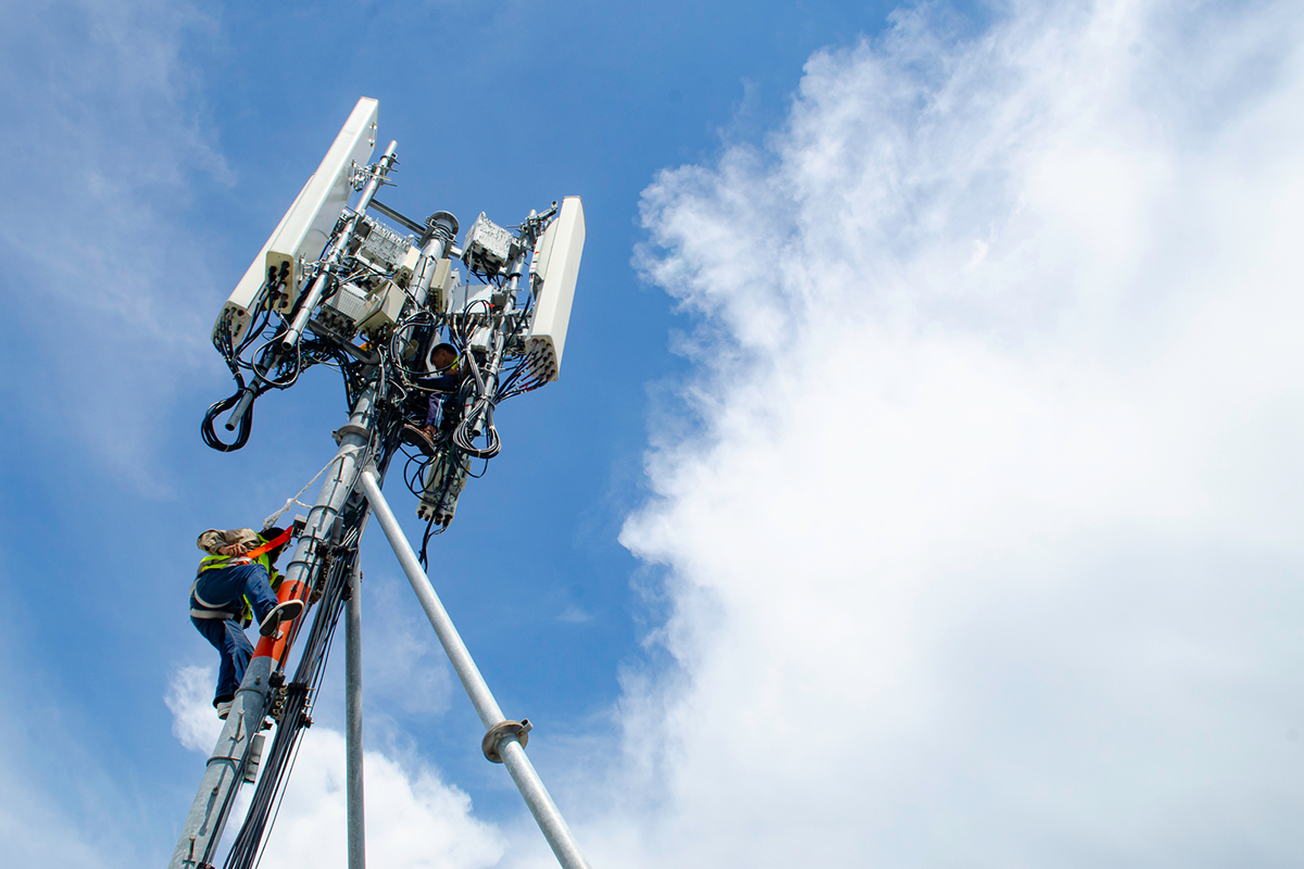 A telecom tower technician works on a cellular tower under a blue sky, simplifying network operations and maintenance. The image represents telecommunications industry infrastructure and field service technology.