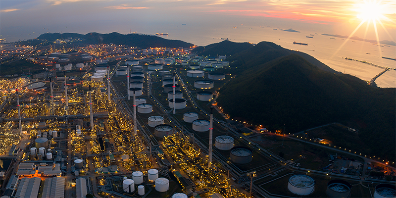 Aerial view of an illuminated industrial oil refinery with storage tanks and chimneys near a coastline at sunset.