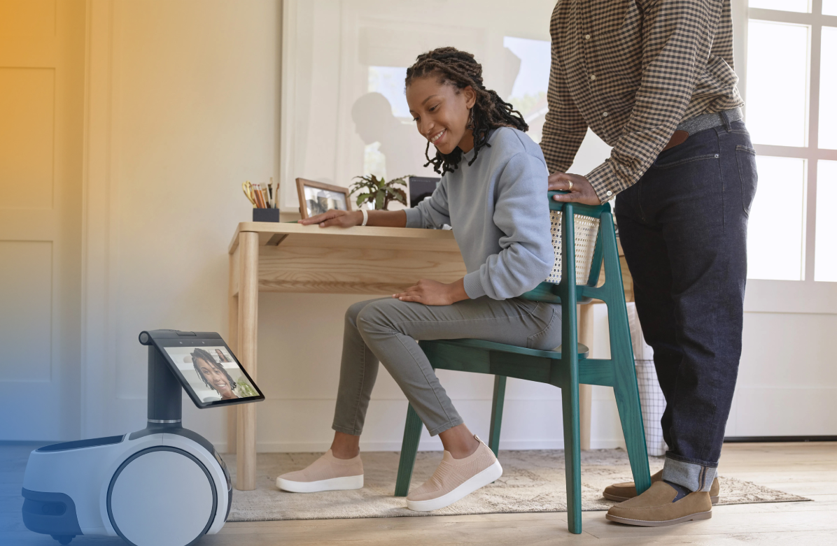 A woman sits at a desk and interacts with a home robot assistant displaying a video call, while another person stands nearby. The setting appears to be a modern home office or living space.