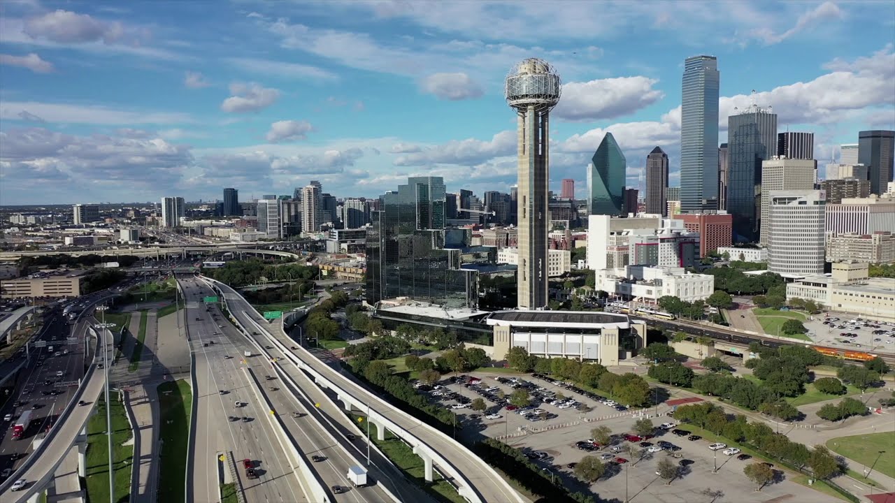 Aerial view of the Dallas skyline featuring the prominent Reunion Tower and surrounding city buildings under a partly cloudy sky.