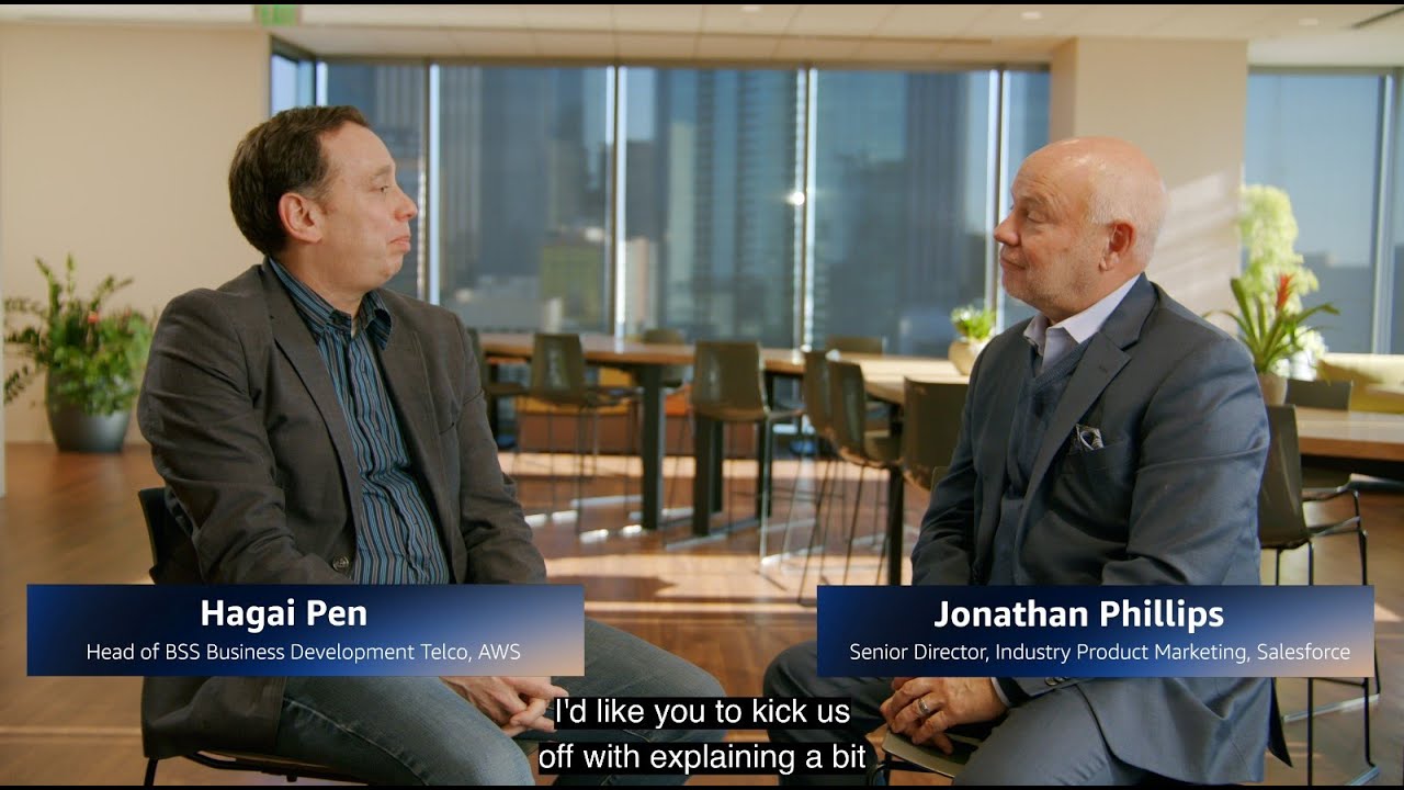 YouTube thumbnail showing a business development interview between two professionals in a modern office setting, featuring on-screen titles for Hagai Pen, Head of BSS Business Development Telco at AWS, and Jonathan Phillips, Senior Director, Industry Product Marketing at Salesforce.