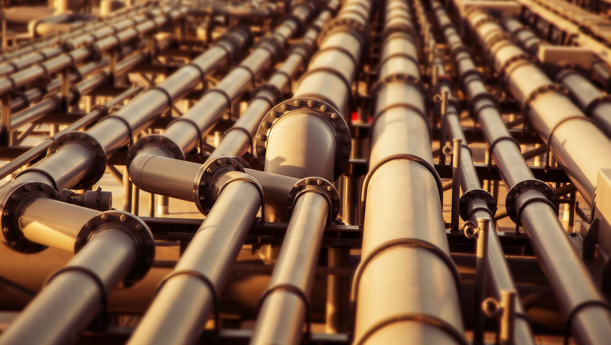 Multiple large industrial pipelines in the oil and gas industry, with a blurred background suggesting depth and infrastructure complexity.