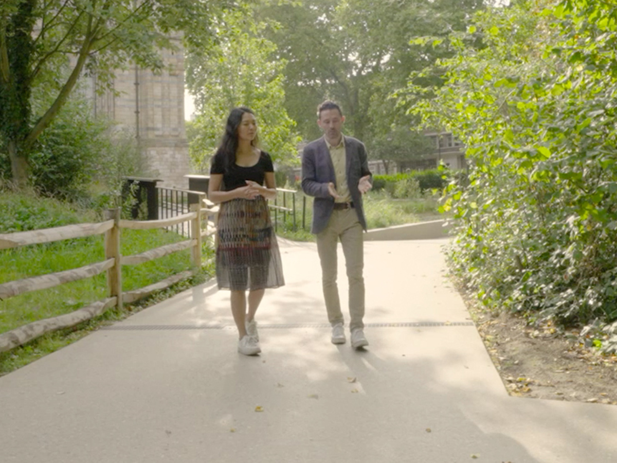 Two people are walking together on a sunny day along a paved garden path, surrounded by greenery and trees.