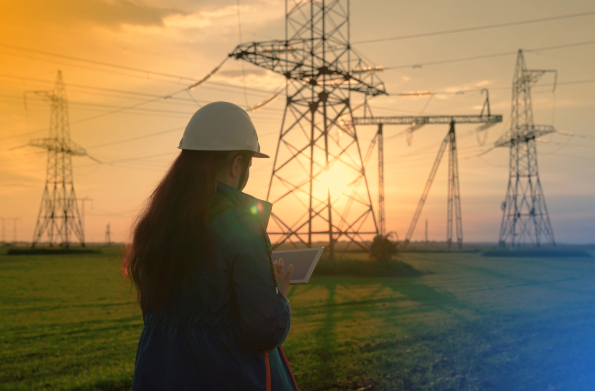 A female engineer wearing a hard hat uses a tablet while inspecting high-voltage power lines and electrical transmission towers at sunset in a rural field.