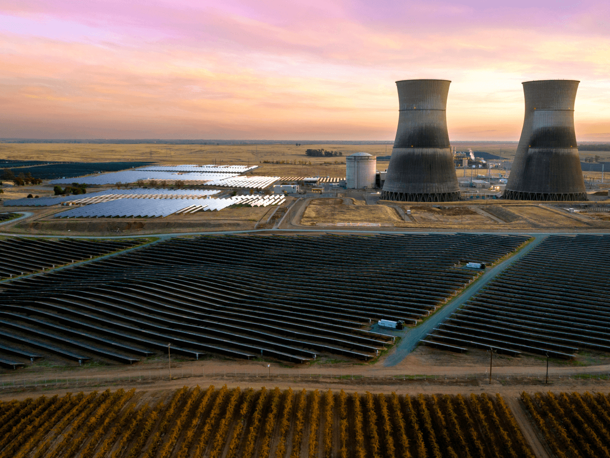Wide view of solar panel arrays in the foreground with power plant cooling towers in the background, all under a colorful sunset sky representing energy and utilities infrastructure.