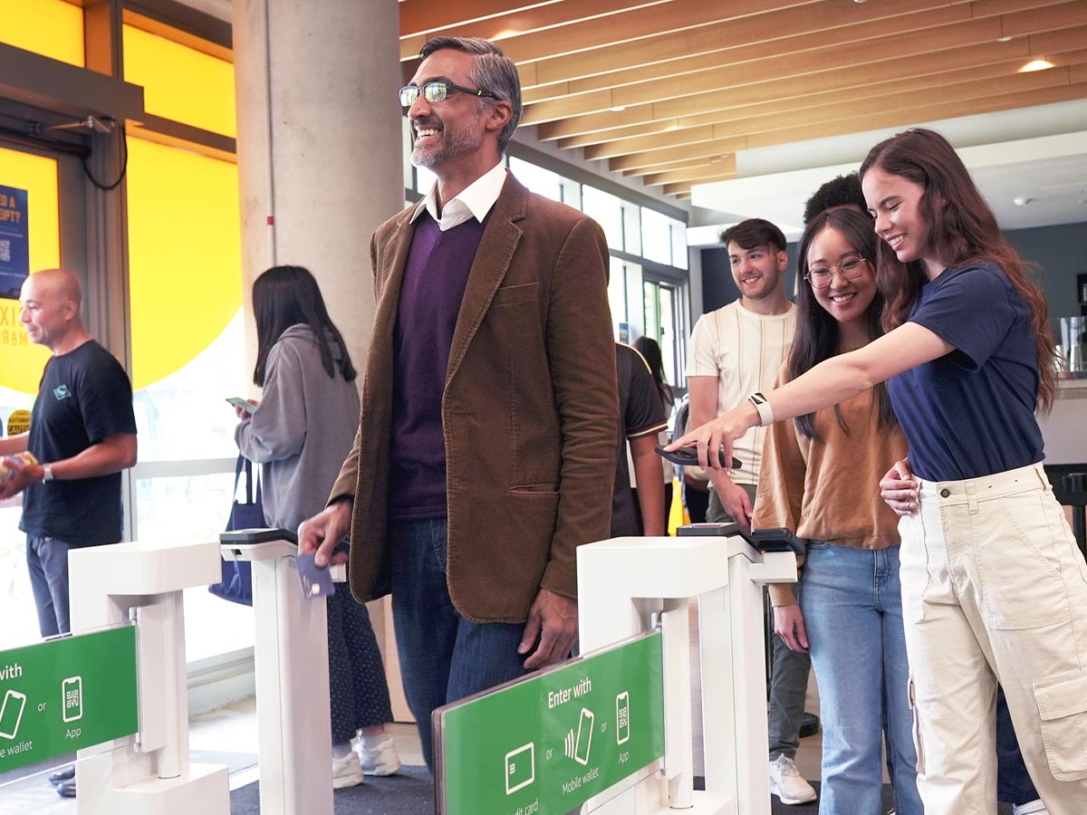 A group of people entering a grab-and-go store by scanning cards and mobile devices at an automated entrance gate.