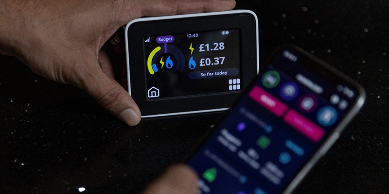 A person holding a smart energy monitor displaying electricity and gas usage costs, with a smartphone showing a related app in the foreground.