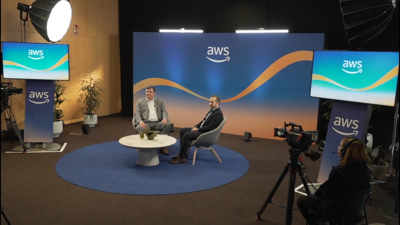 A behind-the-scenes view of an AWS interview studio setup featuring two people seated for an interview with a branded AWS background, lighting equipment, cameras, and display monitors. Ideal for YouTube thumbnails related to AWS interviews, events, or studio production.