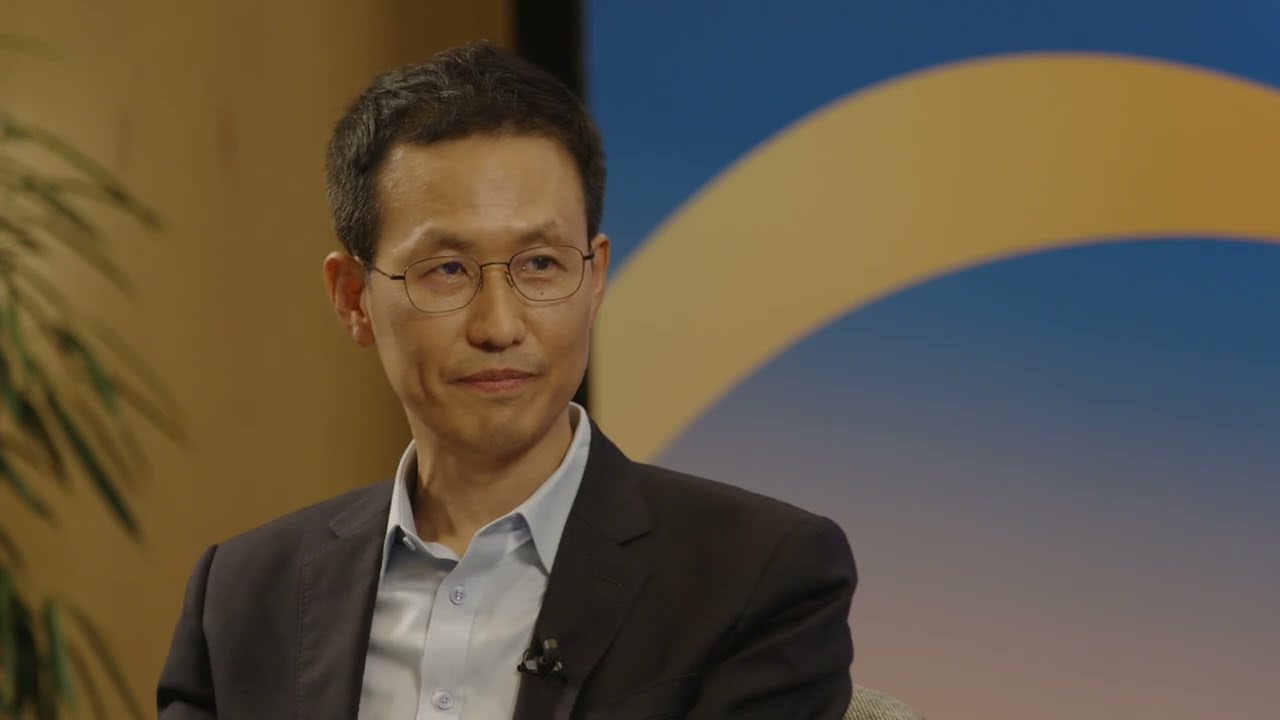 YouTube thumbnail featuring a man in a suit and glasses, seated and being interviewed against a blue and yellow background.