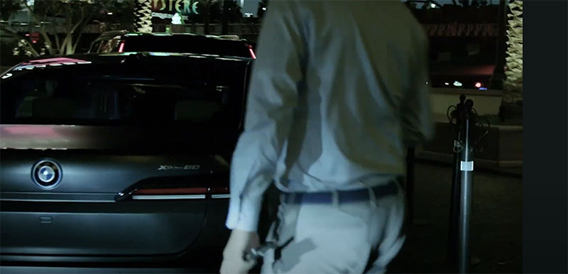 A person is walking toward a parked BMW car at night, with the vehicle's rear and logo visible under artificial lighting in an urban setting.