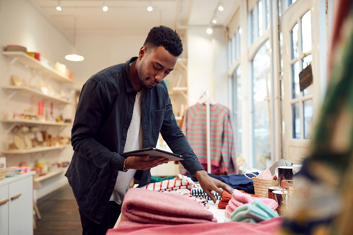 A customer in a retail store browsing clothing displayed on a table while holding a tablet. The store is brightly lit and has shelves and apparel on display.