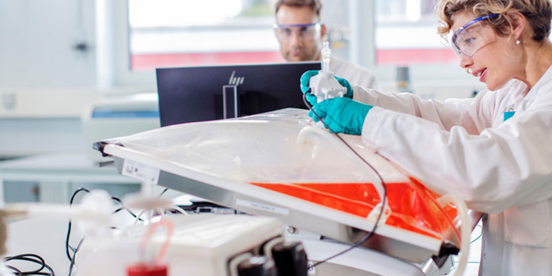 A scientist in a laboratory uses specialized biotechnology equipment while another person works at a computer in the background. Both are wearing lab coats and safety glasses.