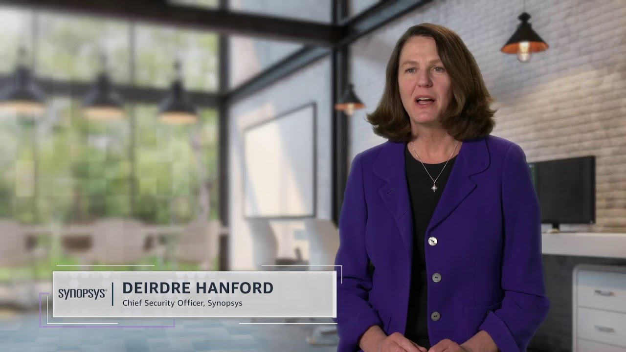 A professional woman in a purple blazer speaks in a modern, bright office setting with large windows and hanging lights. The text overlay identifies her as the Chief Security Officer of Synopsys.