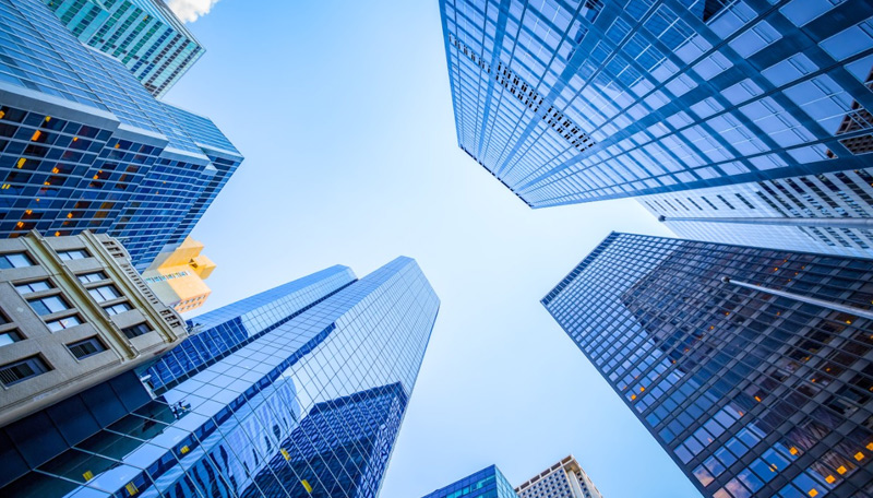 A view looking up at modern skyscrapers and office buildings in a city, with blue sky reflecting off the glass windows.