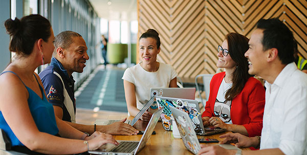 A group of five diverse coworkers collaborating around a desk with laptops in a bright, modern office environment.