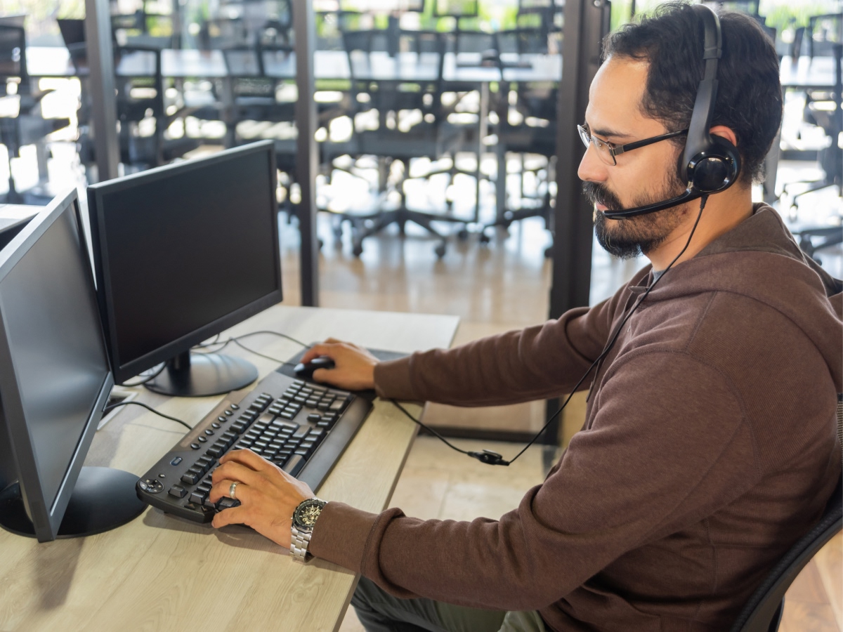 A support agent wearing a headset works at a desk with two monitors