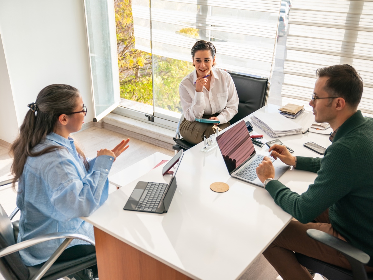 Three colleagues collaborate with laptops around a table in a bright meeting room