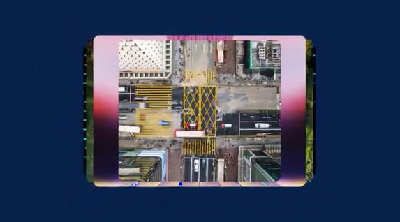 Aerial view of a busy city intersection with yellow crosswalk markings, buses, and surrounding buildings, framed within a smartphone screen.