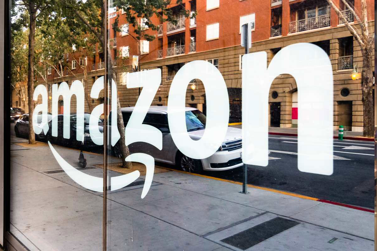 Amazon logo displayed on a large glass window with a city street, parked cars, and buildings reflected in the background.