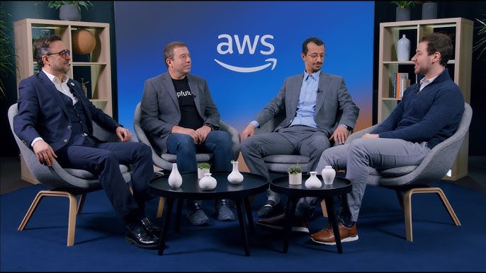 Four men seated in a panel discussion setting with "AWS" branding on a blue backdrop, small tables with decorative vases in front of them.