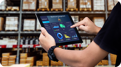 A person uses a tablet displaying a warehouse management dashboard in front of shelves stacked with boxes and inventory in a warehouse storage facility.
