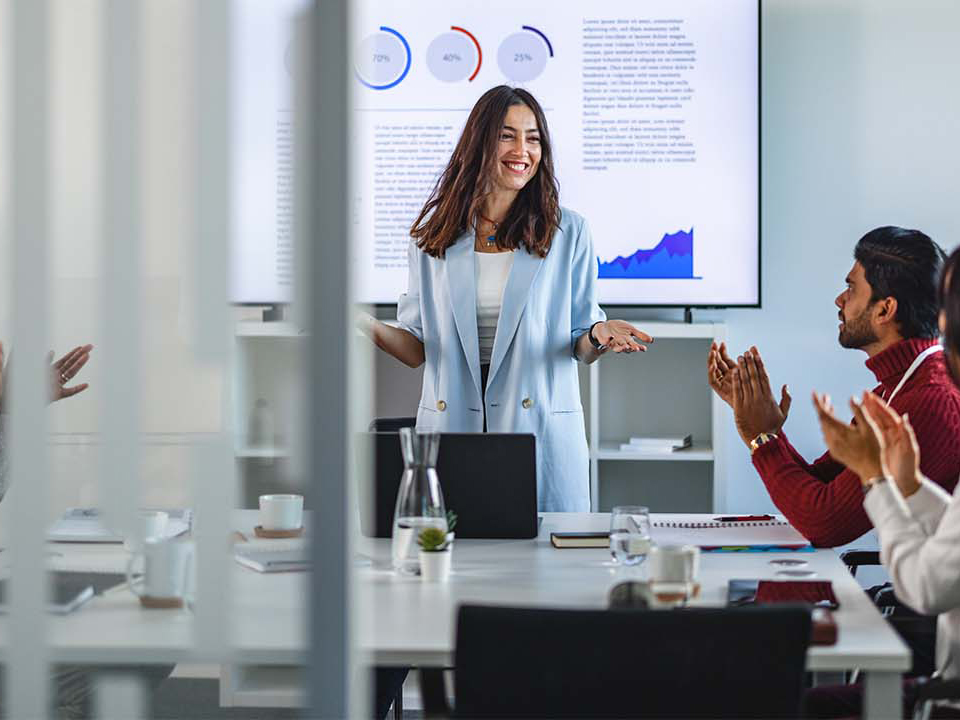 A businesswoman gives a presentation while colleagues sit around a table and applaud. A large screen displaying charts and graphs is visible in the background.