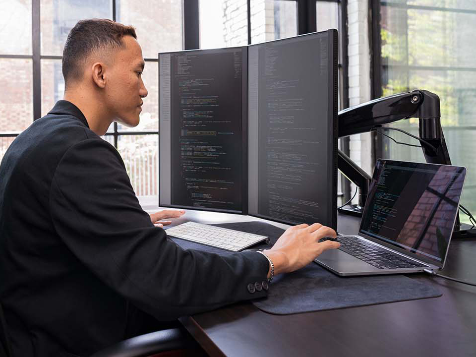 A software engineer working at a desk with three monitors, coding in an office environment. The setup includes two vertical monitors and a laptop, all displaying code, in a modern workspace with large windows in the background.