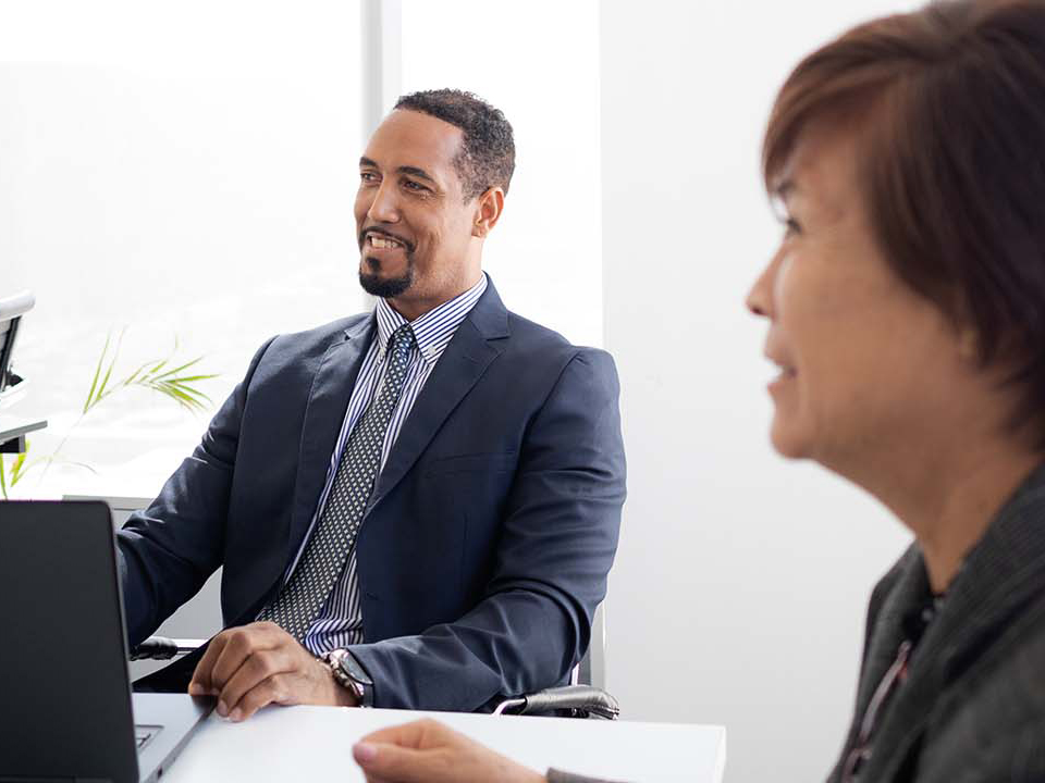 Two business executives smiling and interacting in a modern meeting room, with one working on a laptop.