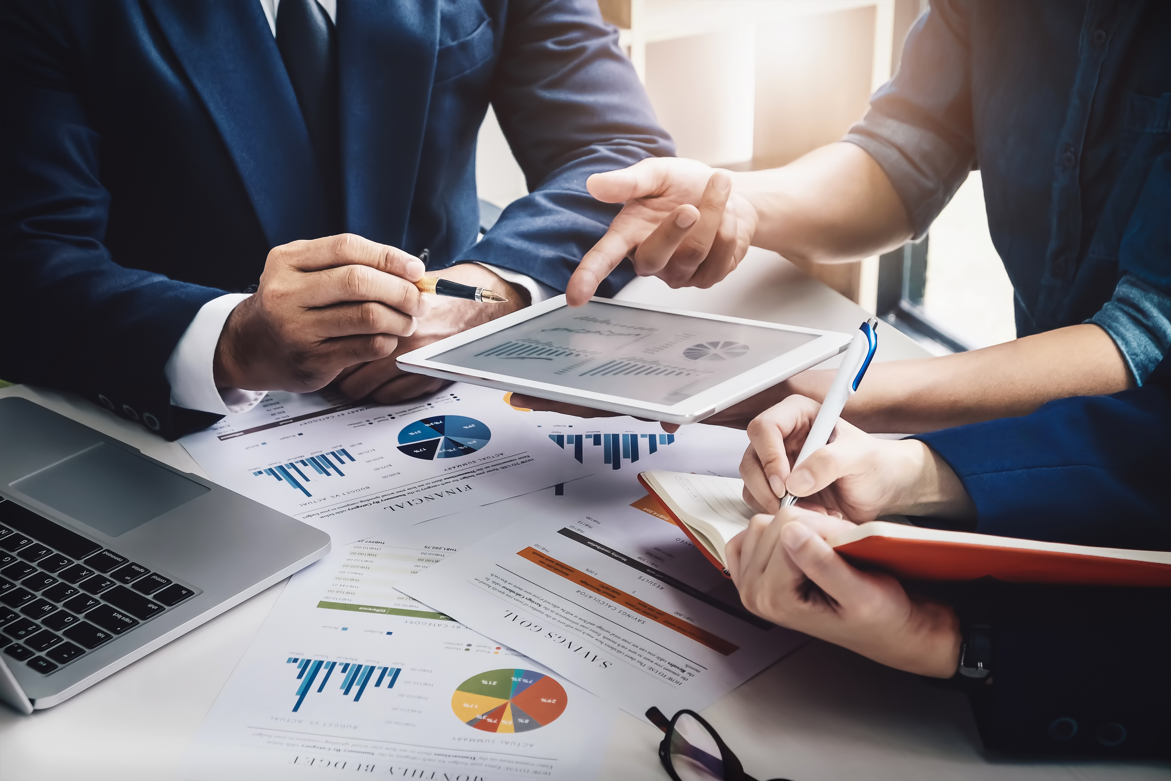 Business professionals review financial charts and graphs on paper and a digital tablet, collaborating on data analysis and taking notes during a meeting.