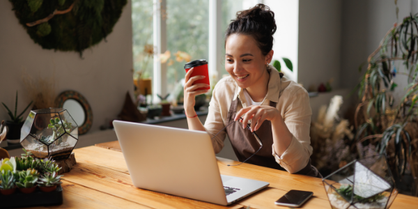 A woman sits at a wooden table with plants, smiling while working on a laptop and holding a coffee cup, suggesting remote work or online learning in a cozy, plant-filled setting.
