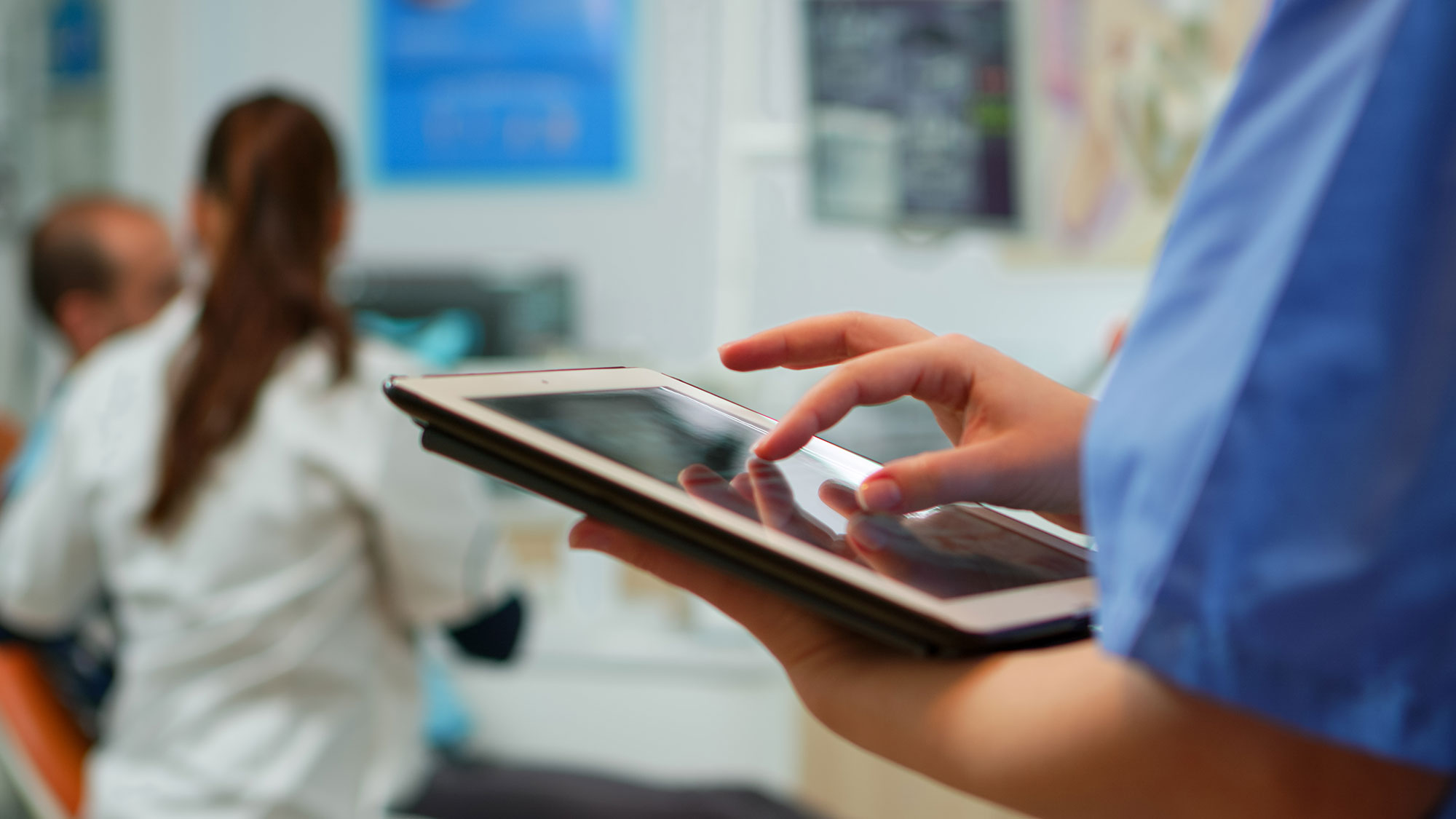 A healthcare professional in scrubs using a digital tablet, with other medical staff and equipment visible in the background of a clinic setting.