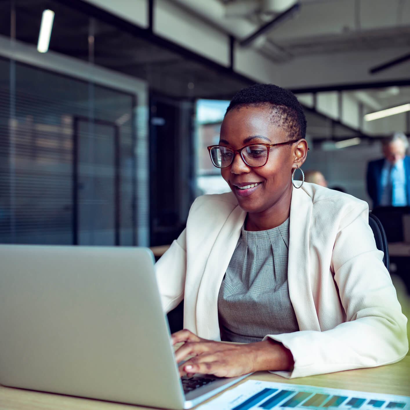 Smiling professional woman wearing glasses and business attire working on a laptop at a desk in a modern office environment, with a document containing graphs in the foreground.