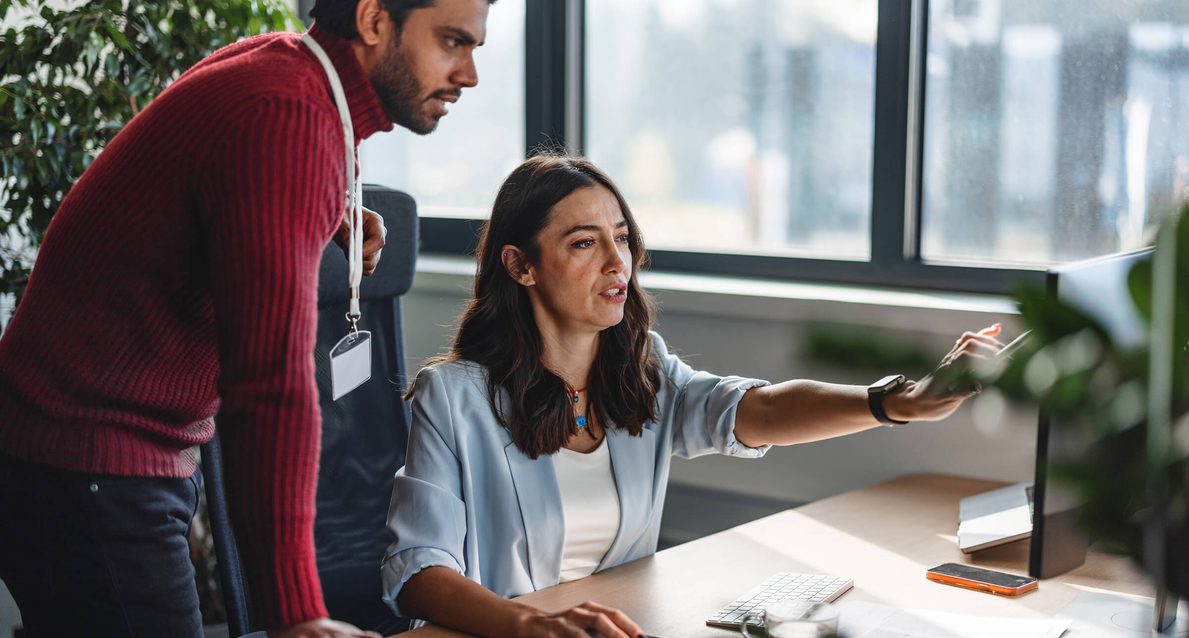 Two colleagues collaborating at a computer desk in a modern office environment. One person is seated and pointing at the computer screen while the other stands beside them, both focused on the task. Large windows and natural light create an open and productive atmosphere.