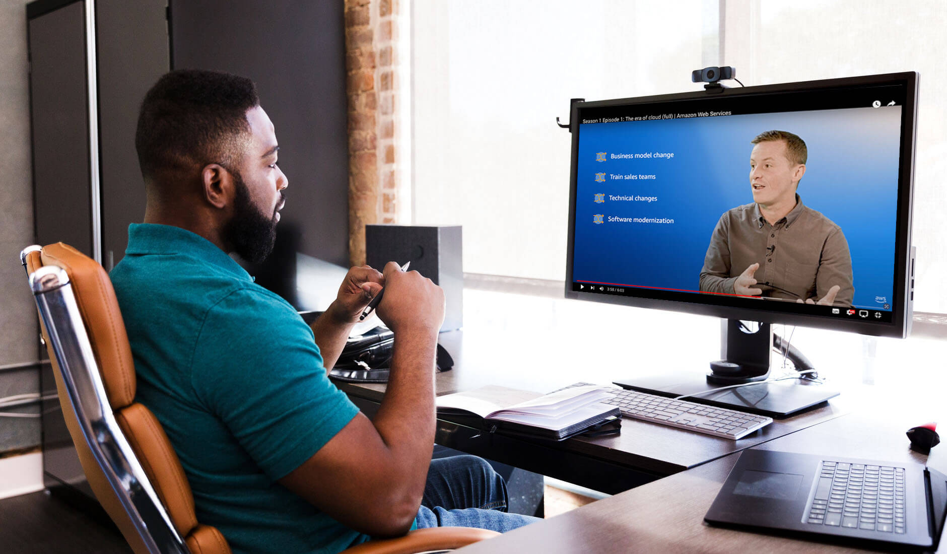 A man sitting at a desk watching a YouTube tech video from Amazon Web Services on his computer monitor, with topics on business model change, training sales teams, technical changes, and software modernization visible on the screen.