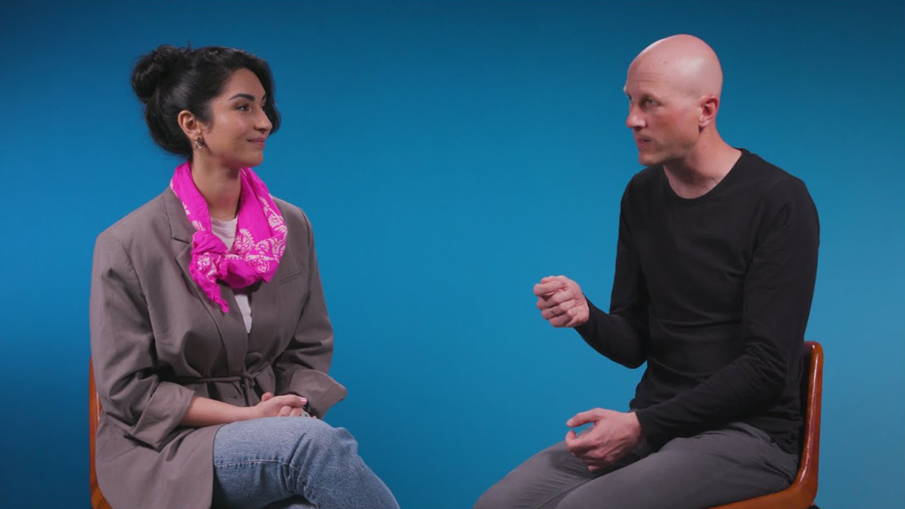 A woman and a man are seated and engaged in a conversation against a solid blue background. The woman is wearing a gray blazer and a bright pink scarf, while the man is dressed in black. Both appear to be in a studio setting.